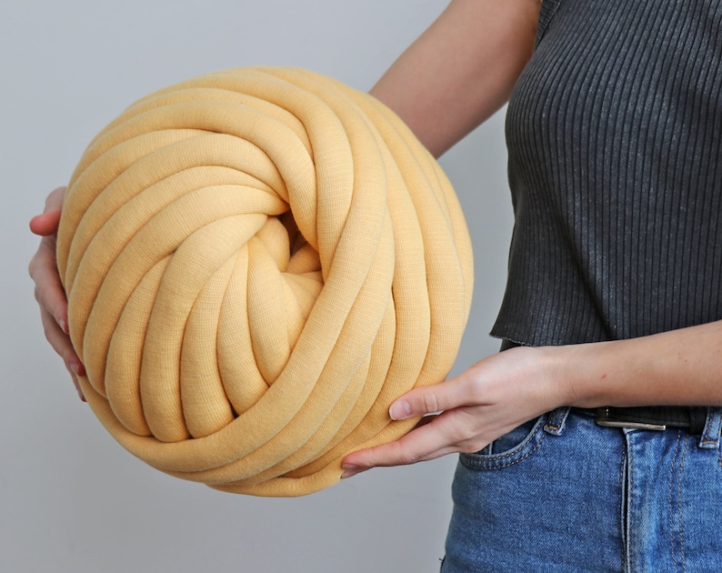 K&ouml;nnte beinhalten: Ein gro&szlig;er, runder Ball aus gelbem Garn, gehalten von einer Person. Das Garn ist dick und scheint aus einem weichen, gestrickten Material zu bestehen. Der Ball wird von einer Person gehalten, was seine Gr&ouml;&szlig;e und Textur zeigt. Ideal f&uuml;r Bastelprojekte.
