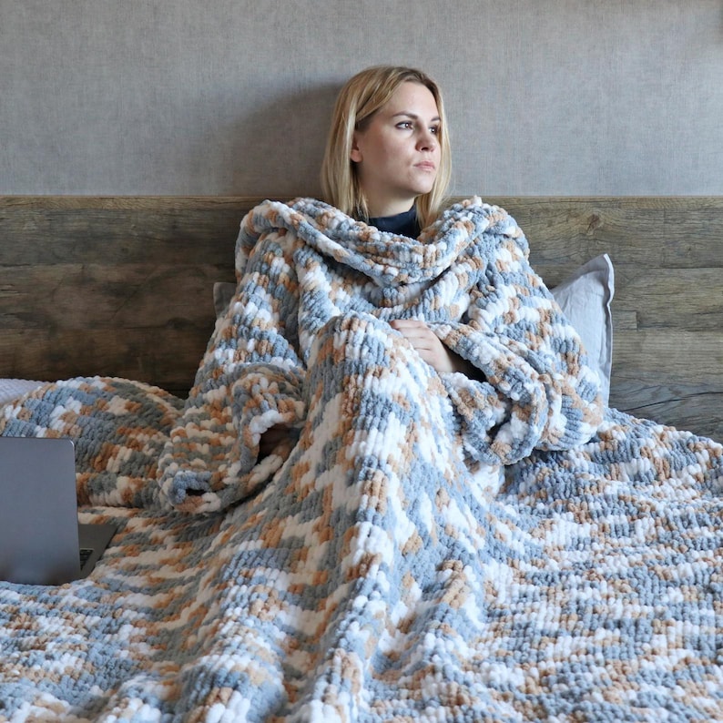 May include: A cozy, hooded blanket in a mix of blue, white, and tan. The blanket is thick and textured, with a soft, plush appearance. The person is sitting on a bed with a laptop.