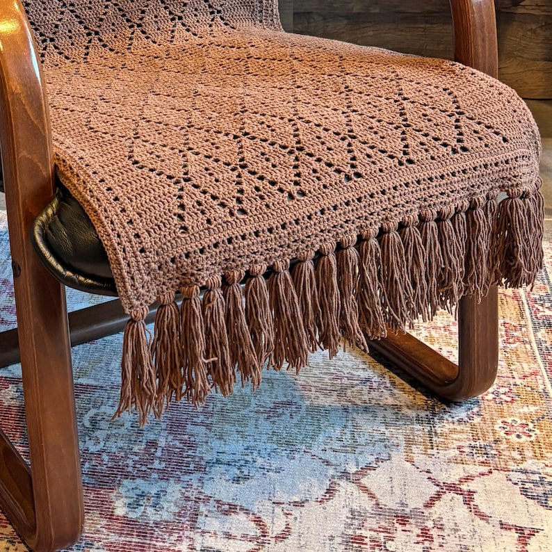 May include: A brown crocheted chair throw with fringe. The throw is on a wooden chair with a brown leather seat. The chair is on a rug with a colorful pattern.