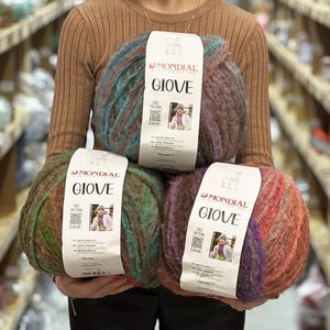 May include: Three large balls of Mondial Giove yarn, each with a white paper label featuring the brand name, a QR code, and a photo of a person wearing a knit garment. The yarn is multicolored with textured, variegated strands in blue, green, and red tones.