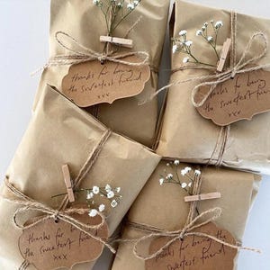 K&ouml;nnte beinhalten: Vier braune Geschenktaschen aus Papier, die mit Schnur gebunden und mit wei&szlig;en Schleierkrautzweigen und handgeschriebenen Etiketten verziert sind, auf denen "Danke, dass du der/die liebste/r Freund/in bist" und "XXX" steht.