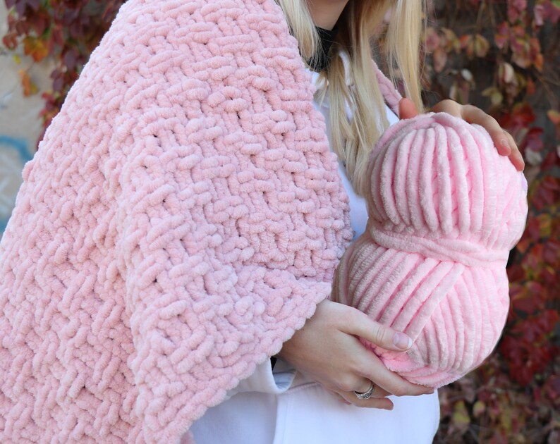 K&ouml;nnte beinhalten: Eine rosa Garnkugel und eine rosa gestrickte Decke. Das Garn ist weich und flauschig, und die Decke ist mit einem dicken Strickmuster gearbeitet.