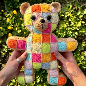 May include: A colourful crocheted teddy bear with a patchwork design. The bear is made of squares in various colours, including pink, orange, yellow, green, and blue. The bear has black eyes and a black nose.