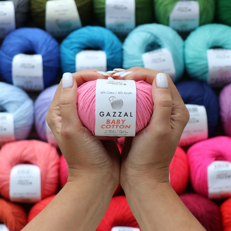 May include: A pink ball of Gazzal Baby Cotton yarn held in hands, with a label indicating 60% cotton and 40% acrylic. The label also shows the weight as 50 g - 1.76 oz. A variety of colorful yarn skeins are in the background.