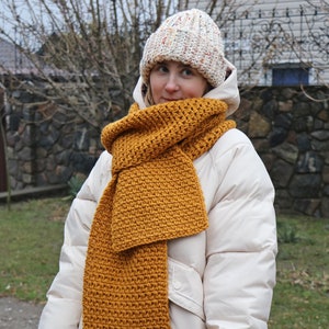 Könnte beinhalten: Eine Frau trägt eine weiße Steppjacke mit einem gelben Häkel-Schal und einer mehrfarbigen Strickmütze.