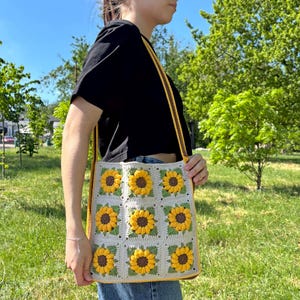 May include: A handmade crochet tote bag featuring a sunflower design. The bag is beige with yellow and brown sunflowers, green leaves, and a yellow strap. The bag is approximately 30 cm wide and 36 cm tall.