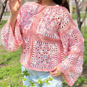 May include: A pink and white crochet top with a square pattern design. The top has long sleeves and a loose fit. The crochet work features intricate floral and geometric patterns, creating a textured and detailed look. The top is made of a soft, lightweight yarn.
