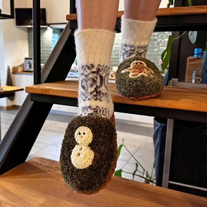 May include: A pair of fuzzy green slippers with embroidered holiday designs. One slipper features a snowman and the other a gingerbread person. The slippers are worn with white socks with a black and white pattern.
