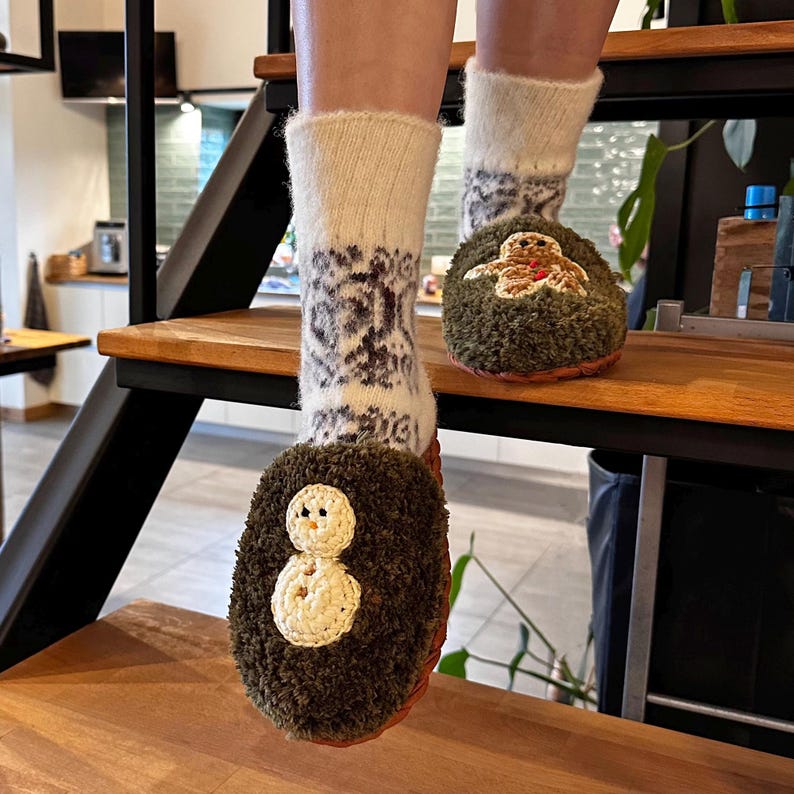 May include: A pair of fuzzy green slippers with embroidered holiday designs. One slipper features a snowman and the other a gingerbread person. The slippers are worn with white socks with a black and white pattern.