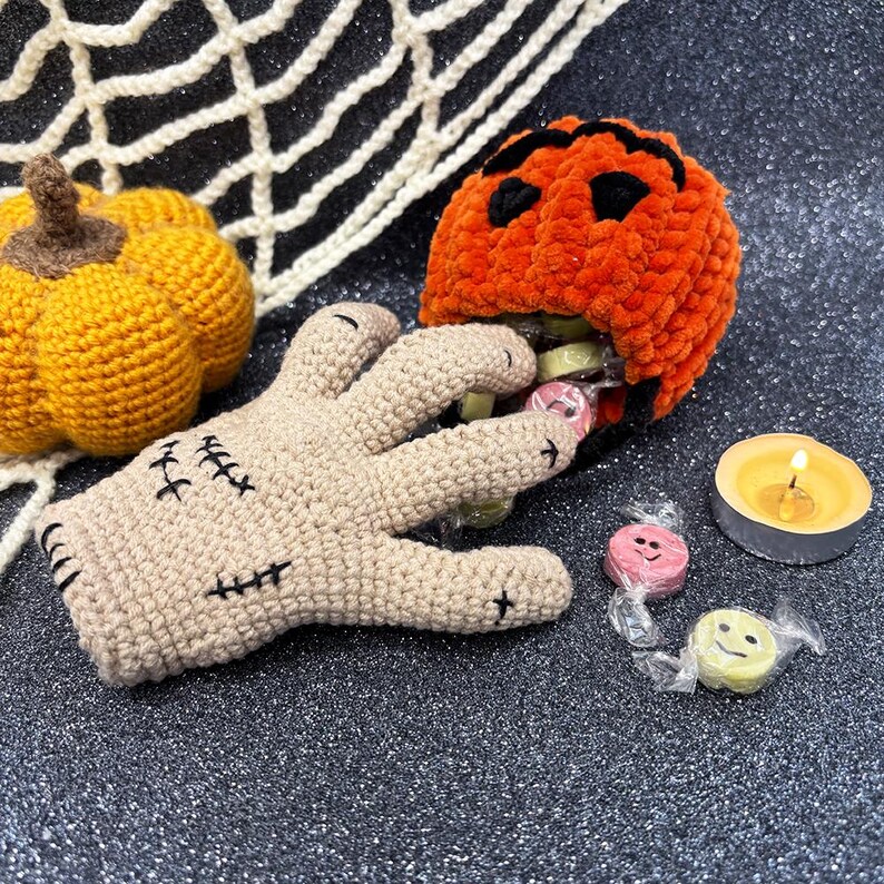 May include: A Halloween-themed crochet display featuring a tan hand with black stitches, an orange pumpkin with a carved face, and a small yellow pumpkin. The hand holds wrapped candies, with a lit candle nearby.