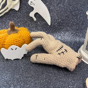 May include: A tan crochet hand with black stitched details, next to a small orange crochet pumpkin and white foam ghost decorations. The Halloween-themed items are on a dark, glittery surface.
