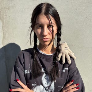 May include: A dark gray sweatshirt with a graphic print and the word "BLOODE" is worn. A small, beige crocheted glove with stitched details rests on the shoulder. The person has dark hair in braids and red nail polish.
