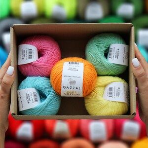 May include: A cardboard box filled with colorful yarn skeins. The yarn is in shades of pink, orange, teal, green, and yellow. Each skein has a label. The yarn is labeled "GAZZAL BABY COTTON".