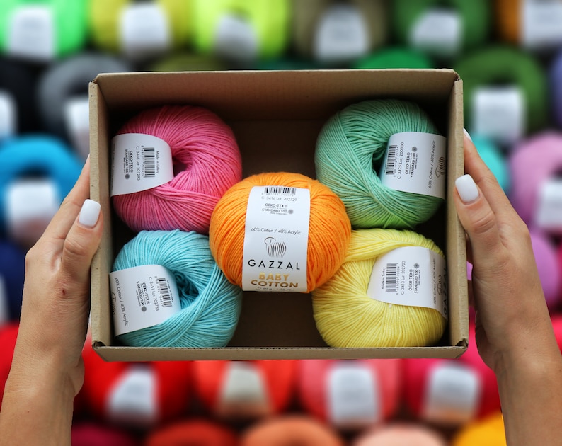 May include: A cardboard box filled with colorful yarn skeins. The yarn is in shades of pink, orange, teal, green, and yellow. Each skein has a label. The yarn is labeled "GAZZAL BABY COTTON".