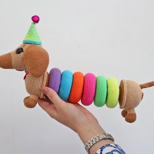 May include: A crocheted dachshund toy wearing a green and white party hat with a pink bell. The dachshund's body is made of brown yarn and has a rainbow of colorful crocheted rings around its middle.