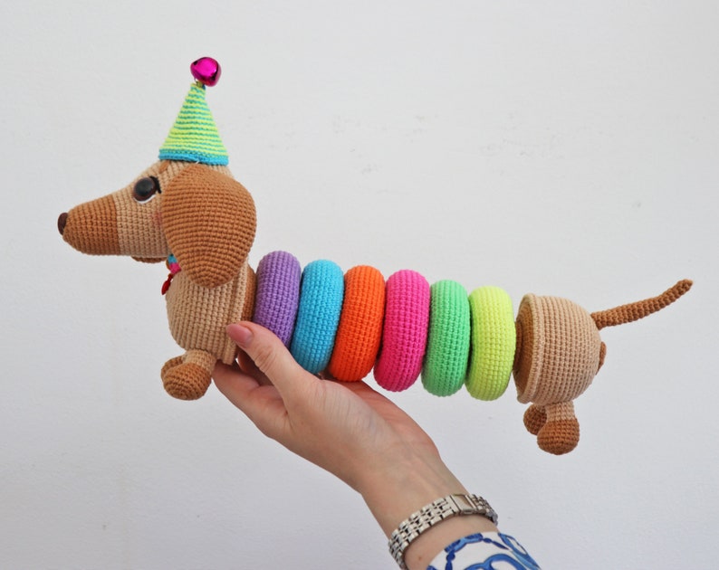May include: A crocheted dachshund toy wearing a green and white party hat with a pink bell. The dachshund's body is made of brown yarn and has a rainbow of colorful crocheted rings around its middle.