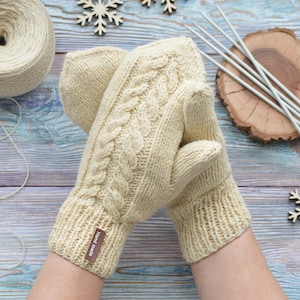 May include: A pair of cream-colored knitted mittens with a cable knit pattern. The mittens have a brown leather tag that says "Handmade".