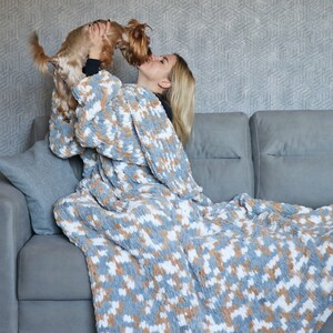 May include: A woman wearing a grey and white patterned blanket as a robe is sitting on a grey sofa. She is holding a small brown and white dog in her arms. The dog is licking her face.