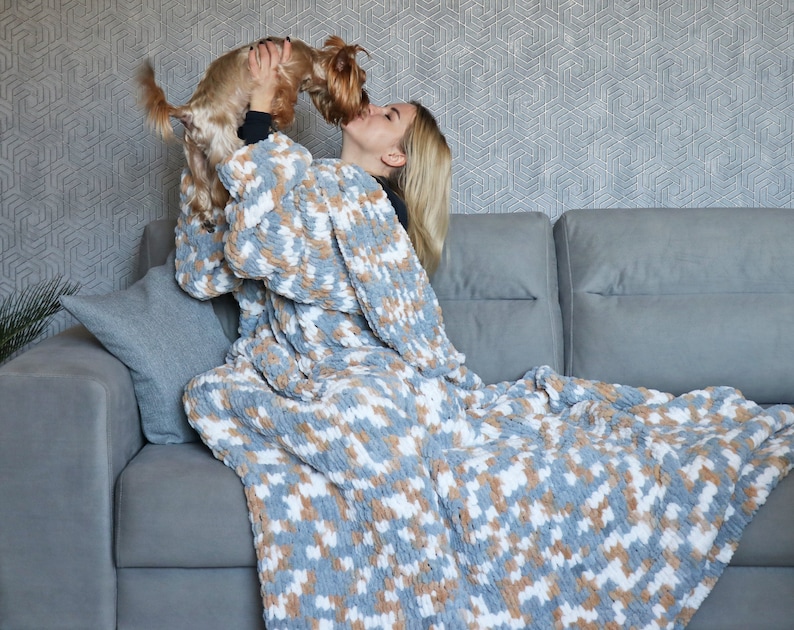 May include: A woman wearing a grey and white patterned blanket as a robe is sitting on a grey sofa. She is holding a small brown and white dog in her arms. The dog is licking her face.