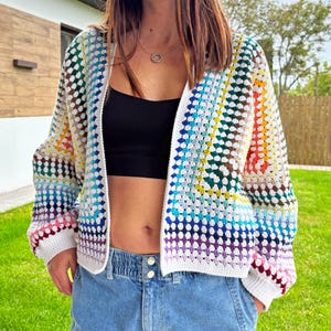 Peut inclure: Un cardigan en crochet ouvert, aux couleurs vives. Le vêtement présente un arc-en-ciel de couleurs, notamment le rouge, l'orange, le jaune, le vert, le bleu et le violet, disposées en motif carré. Les bords sont finis avec une bordure blanche. Le cardigan est porté sur un haut noir et un jean bleu.