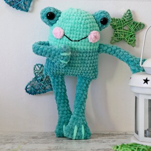May include: A crocheted green frog toy with pink cheeks and large black eyes. The frog is standing on a white surface.