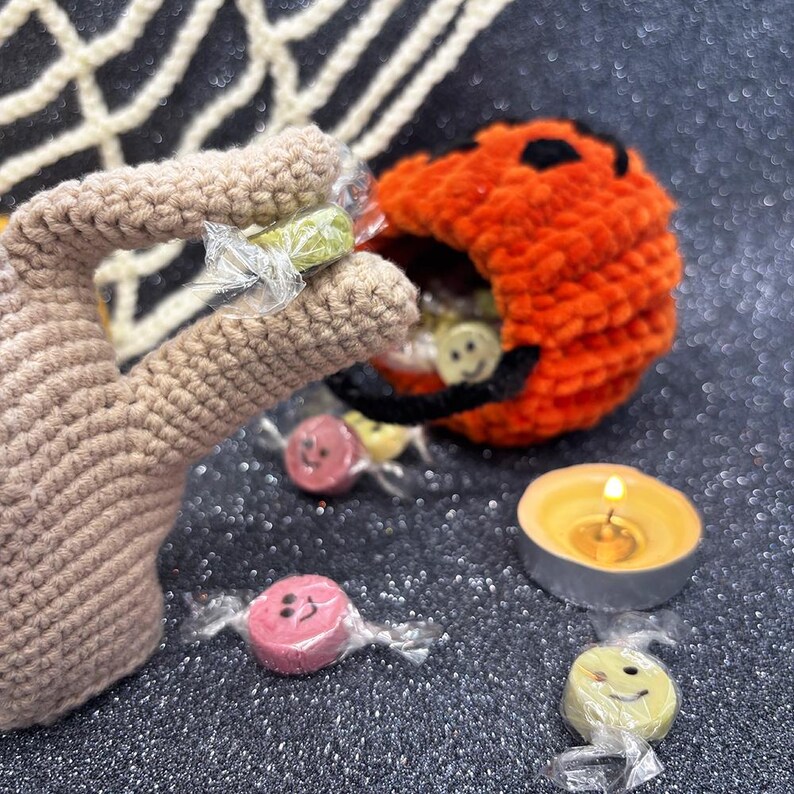 May include: A crocheted pumpkin candy holder filled with candies. The pumpkin is orange with a black face. The candies are wrapped in clear cellophane and have smiley faces. A lit candle and a crocheted hand are also in the image.