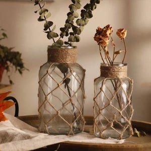 K&ouml;nnte beinhalten: Zwei klare Glasvasen mit einem Seilnetz-Design. Die Vasen sind mit getrockneten Blumen und Gr&uuml;n gef&uuml;llt. Die Vasen stehen auf einer Holzoberfl&auml;che.