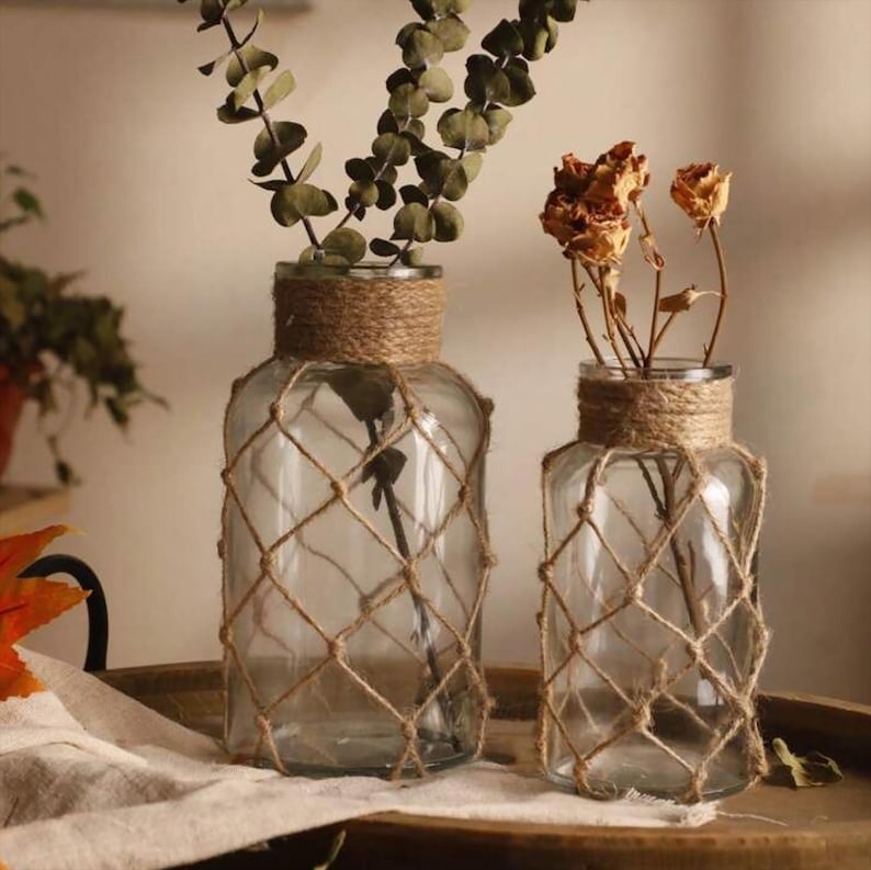 K&ouml;nnte beinhalten: Zwei klare Glasvasen mit einem Seilnetz-Design. Die Vasen sind mit getrockneten Blumen und Gr&uuml;n gef&uuml;llt. Die Vasen stehen auf einer Holzoberfl&auml;che.