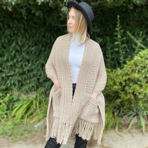 Może przedstawiać: Beżowy szal zrobiony na szydełku z frędzlami i dwoma kieszeniami. Szal nosi osoba ubrana w biały golf i czarne spodnie. Osoba nosi czarny kapelusz.