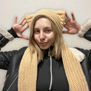 Könnte beinhalten: Eine Frau trägt eine beige gestrickte Mütze mit Katzenohren und einen passenden Schal. Mütze und Schal sind gehäkelt und haben ein strukturiertes Muster.