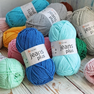 Puede incluir: Surtido de ovillos de lana en varios colores, incluyendo azul, verde, rosa, amarillo y gris. Los ovillos de lana tienen etiquetas que dicen "YarnArt jeans plus".