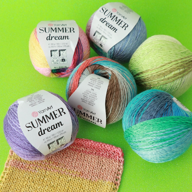 May include: Five balls of yarn in various colors, including purple, green, blue, brown, and pink. Each ball has a label that reads "YarnArt Summer Dream" and includes the weight and yardage of the yarn. The yarn is arranged on a green surface.