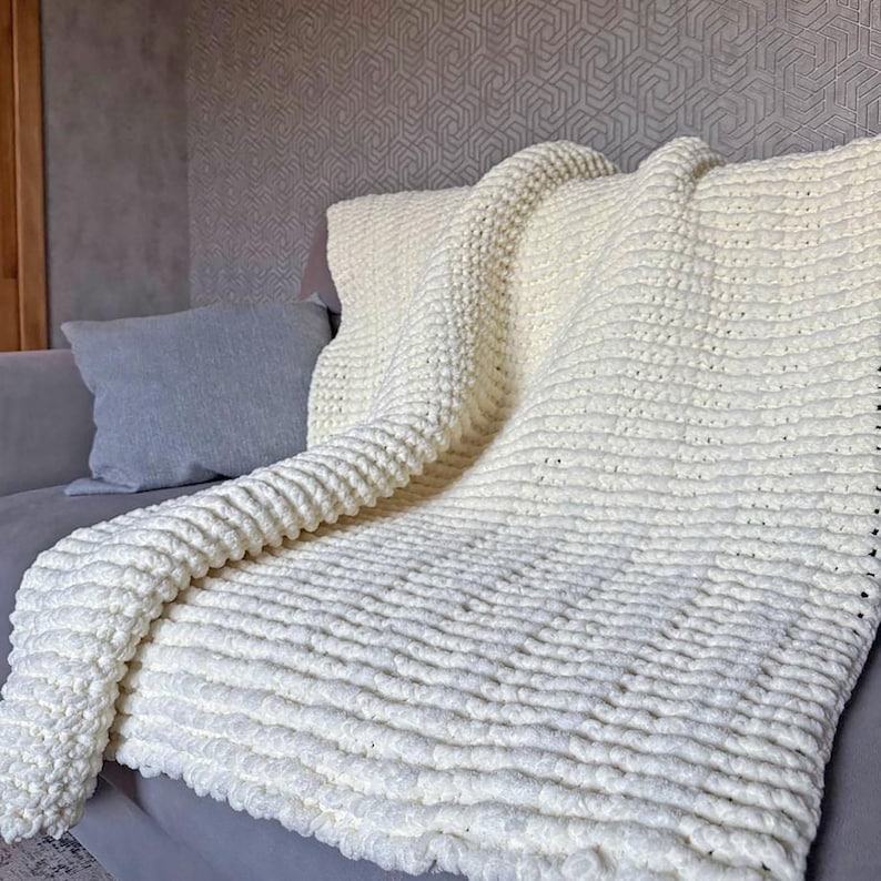 May include: A cream-colored, chunky knit blanket draped over a gray sofa. The blanket has a textured, ribbed pattern and appears soft and cozy. A gray pillow rests on the sofa, complementing the blanket's neutral tone. The background features a patterned wall.