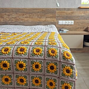 K&ouml;nnte beinhalten: Eine handgefertigte H&auml;keldecke mit einem sich wiederholenden Sonnenblumenmuster. Die Decke hat einen grauen Hintergrund mit braunen R&auml;ndern und gr&uuml;nen Bl&auml;ttern. Die Sonnenblumen sind gelb mit braunen Zentren. Die Decke liegt auf einem Bett.
