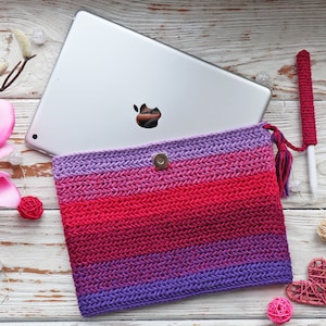 May include: A handmade crocheted tablet sleeve in a gradient of pink and purple shades, with a magnetic clasp. A matching stylus holder and a tablet are also visible. The sleeve rests on a light wooden surface.