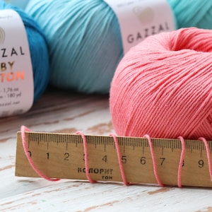 May include: Close-up of colorful yarn skeins and a wooden ruler. The yarn is in shades of pink, teal, and blue. The ruler measures the pink yarn strands. The yarn label reads "GAZZAL BABY COTTON".