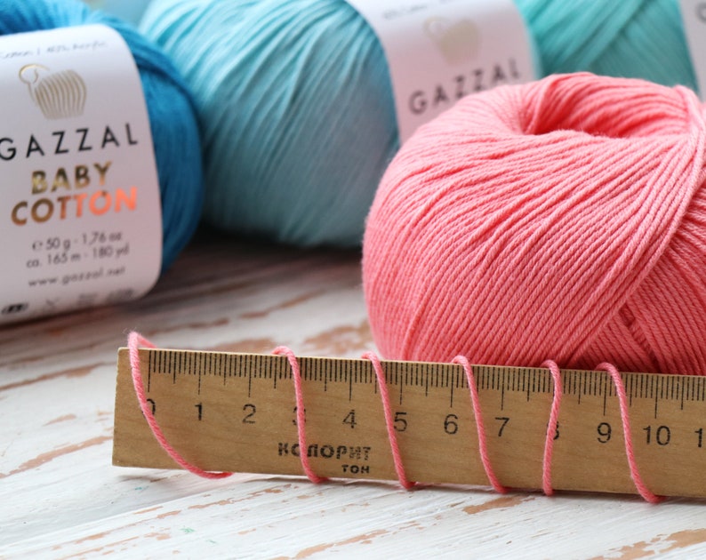 May include: Close-up of colorful yarn skeins and a wooden ruler. The yarn is in shades of pink, teal, and blue. The ruler measures the pink yarn strands. The yarn label reads "GAZZAL BABY COTTON".