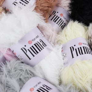 May include: A selection of YarnArt Piuma yarn skeins in shades of white, cream, brown, and black. The yarn has a soft, fluffy texture. Each skein has a white label with the word "Piuma" in large black letters.