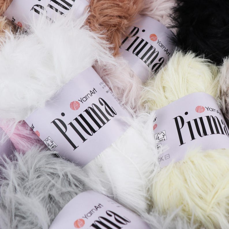May include: Close-up of several skeins of YarnArt Piuma yarn in various colors, including white, cream, brown, and black. The yarn has a fluffy, feathery texture. The labels are white with the word "Piuma" in large black letters.
