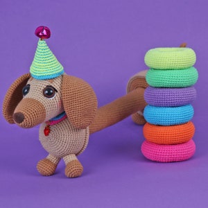 May include: A brown crocheted dachshund wearing a green and yellow party hat with a pink bell on top. The dachshund is standing on a purple surface with a stack of colorful crocheted rings.
