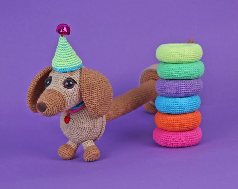 May include: A brown crocheted dachshund wearing a green and yellow party hat with a pink bell on top. The dachshund is standing on a purple surface with a stack of colorful crocheted rings.