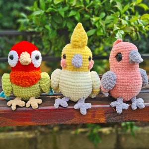 May include: Three crocheted bird plushies in different colors. The bird on the left is red and green, the middle bird is yellow and white, and the bird on the right is pink and gray.