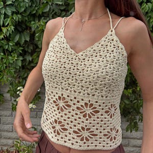 May include: Cream-coloured crochet halter top with a V-neckline and floral design. The top features a halter strap and a detailed openwork pattern. The handmade top is a summer garment.