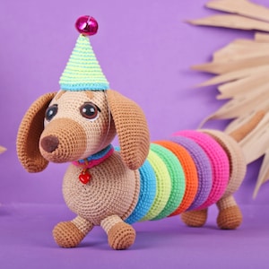 May include: A crocheted dachshund wearing a party hat with a pink pom-pom. The dachshund is brown and has a rainbow-colored tail.