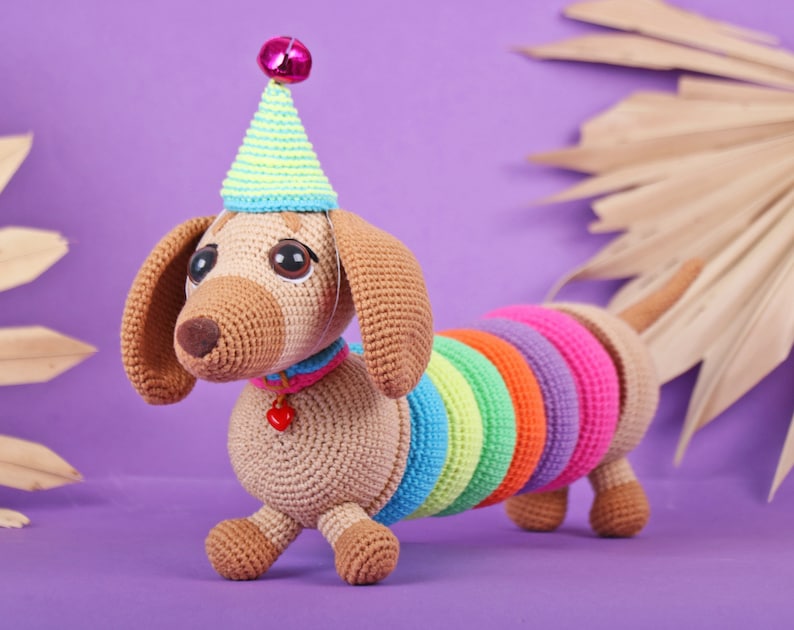 May include: A crocheted dachshund wearing a party hat with a pink pom-pom. The dachshund is brown and has a rainbow-colored tail.