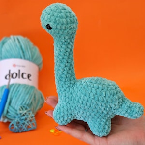 Puede incluir: Un juguete de dinosaurio tejido a ganchillo de color turquesa con un cuello largo y patas pequeñas. El juguete de peluche se sostiene en una mano sobre un fondo naranja. Un ovillo de lana turquesa y un ganchillo son visibles en el fondo.