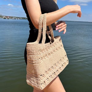 May include: A beige crochet tote bag with two handles. The bag features a textured pattern and is held against a backdrop of water and a blue sky. The bag is a neutral color, suitable for various occasions.