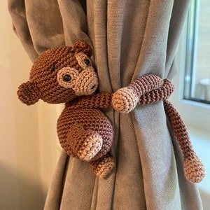 May include: A brown crocheted monkey toy with a long tail used as a curtain tieback. The monkey is holding onto a beige curtain.