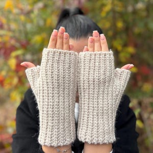 Puede incluir: Un par de guantes sin dedos beige, tejidos con un patrón de canalé. Los guantes están hechos de un material suave y cálido, ideal para mantener las manos abrigadas. Los guantes se muestran sobre un fondo borroso de follaje otoñal.