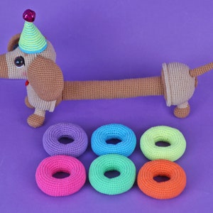 May include: A crocheted dachshund wearing a party hat and holding six colorful crocheted rings.